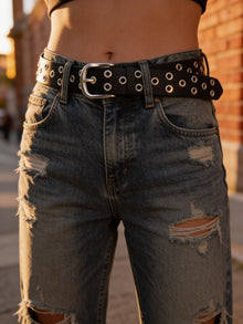 Cinturón negro con ojales metálicos de La tiendita de Carlita sobre jeans desgastados, estilo casual y moderno.